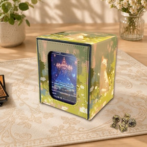 Puede incluir: Una caja cuadrada para guardar cartas con una ilustración de un gato sobre un fondo verde y amarillo. La caja tiene una ventana que muestra una carta con texto. Los dados son visibles en primer plano.