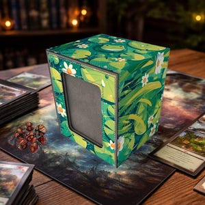 May include: A green and white card storage box with a frog and lily pad design. The box has a rectangular window and is surrounded by a white stitched border. Dice and cards are visible on the table.