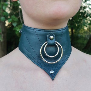 Könnte beinhalten: Ein dunkelgrüner Leder-Choker mit einem silbernen Ring und einem silbernen Niet in der Mitte. Der Choker ist mit Nähten verziert und hat eine spitze Form.
