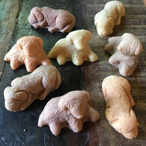 May include: Eight handmade soap bars shaped like dogs in various poses. The soaps are in shades of brown and beige.