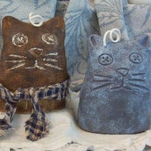 May include: Two handmade ceramic cat candle holders. One is brown with a blue and white plaid scarf and the other is gray. Both have painted details and a white wick.