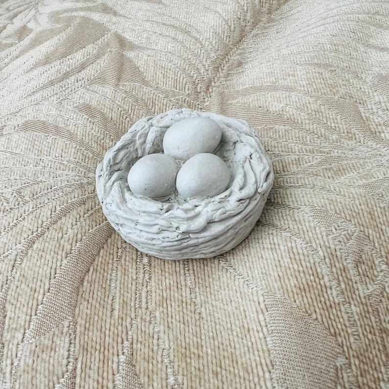 Op de afbeelding: Een wit keramisch vogelnest met drie witte eieren erin. Het nest ligt op een beige, getextureerd oppervlak.