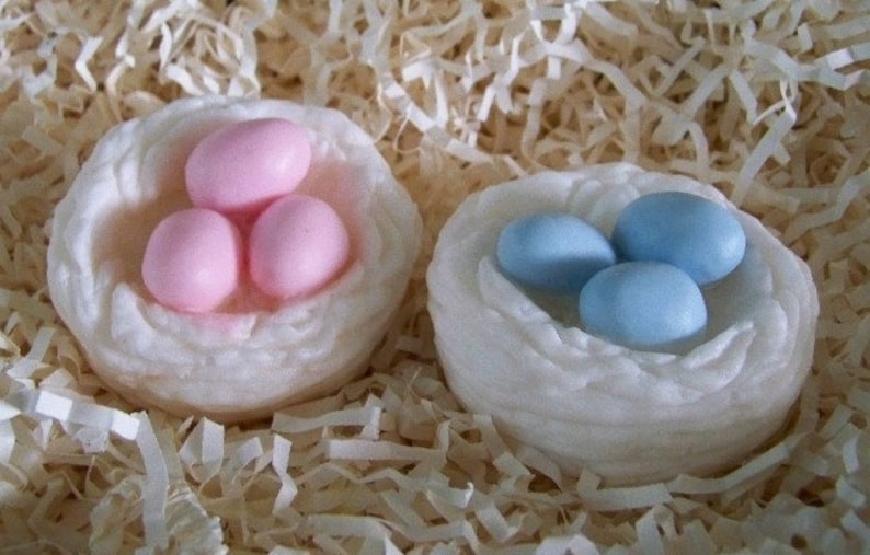 Op de afbeelding: Twee witte nesten, een met drie roze eieren en de andere met drie blauwe eieren. De nesten zijn gemaakt van een zacht, pluche materiaal en liggen op een bed van gescheurd papier.