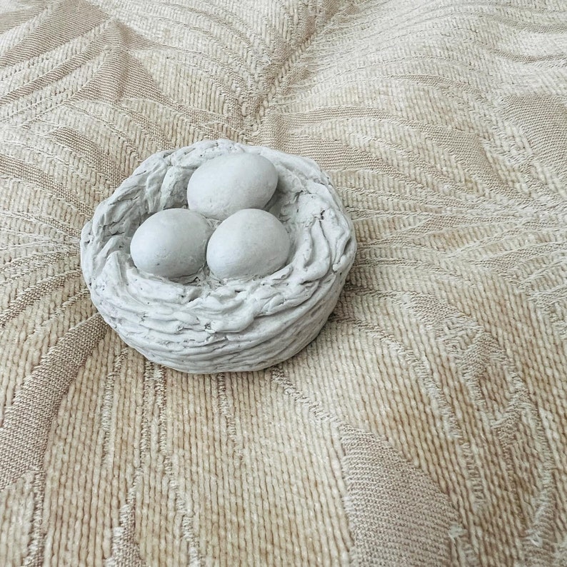Op de afbeelding: Een wit keramisch vogelnest met drie witte eieren erin. Het nest ligt op een beige, getextureerd oppervlak.