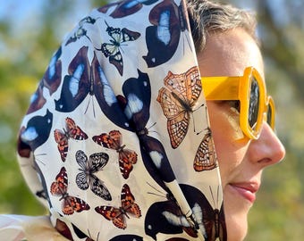 May include: A cream-colored headscarf featuring a repeating pattern of butterflies in shades of brown, black, and blue. The scarf is worn on the head and is visible from the side. The person is wearing yellow sunglasses.