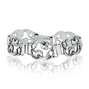 May include: A silver bracelet with an intricate, openwork pattern. The design incorporates floral and scroll elements, linked by rectangular sections. The clasp is a simple bar style. The bracelet is shown against a white backdrop.