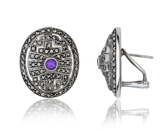 May include: A pair of silver earrings with a purple gemstone in the center of each earring. The earrings are surrounded by a design of small black stones.