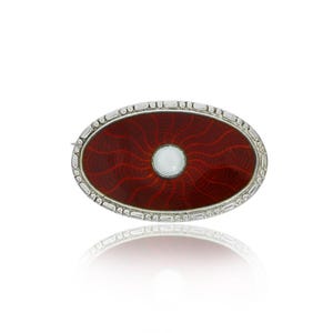 May include: An oval brooch featuring a red enamel design with a white pearl at its centre. The brooch is bordered by a silver frame with a decorative pattern.
