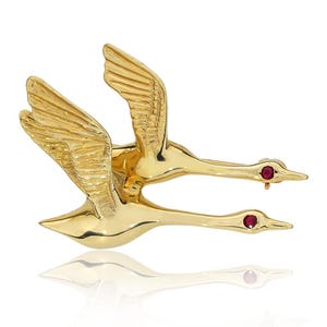 Op de afbeelding: Een broche van goudkleurig metaal met twee gestileerde vogels in vlucht met robijnrode accenten.