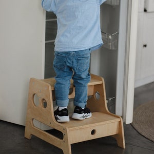 May include: A young child standing on a wooden step stool reaching for something inside a white fridge. The child is wearing a light blue button-down shirt, blue jeans, and black trainers.