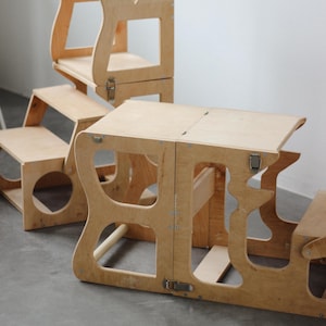 May include: A set of unfinished wooden children's furniture, including a table and chairs. The table has a hinged top and the chairs are stackable. The furniture is made of light-colored wood and has a simple, modern design. The set is designed for children.