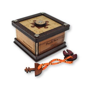 May include: A wooden puzzle box with a square shape and a light wood top featuring a jigsaw puzzle design. The box has dark wood trim and metal accents. The text "Puzzle Box 04" is visible. A wooden key with an orange and yellow rope is also present.