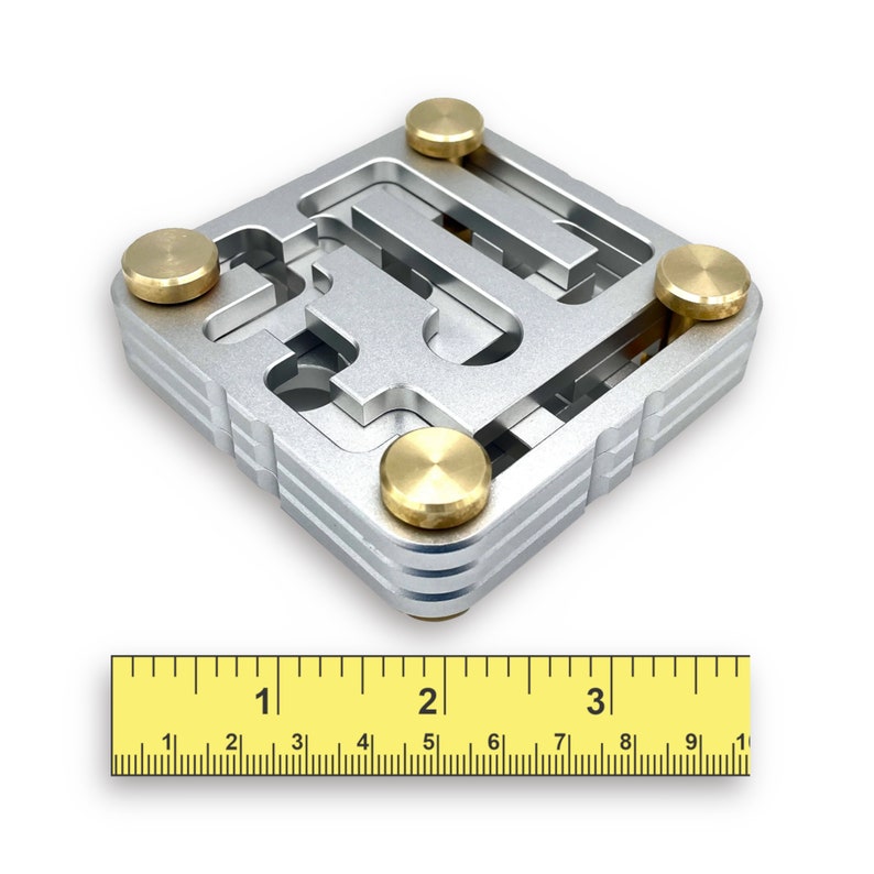 May include: A silver-colored puzzle with a maze-like design, featuring four brass-colored circular knobs. The puzzle is square-shaped and has a ruler with inch measurements at the bottom. The puzzle measures approximately 3 inches in length.