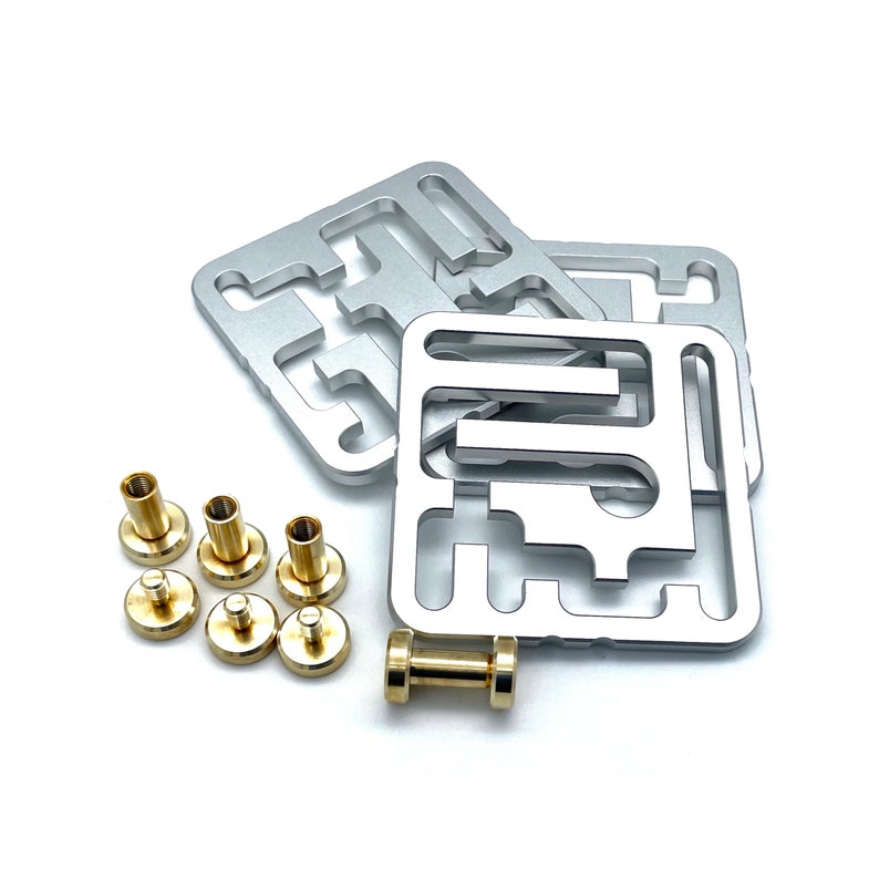 May include: A collection of silver metal square components with intricate cut-out designs, alongside several brass-colored cylindrical fasteners and a small brass dumbbell-shaped object. The items are arranged on a white surface.