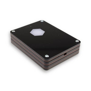 May include: A black rectangular box with a hexagonal cutout in the centre. The box is made of layered acrylic and has a wood-like finish. The box is secured with screws on the edges.
