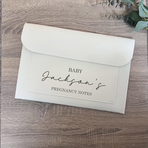 May include: A cream-coloured envelope-style organiser with a flap closure. The front features a rectangular patch with the text "BABY Jackson's PREGNANCY NOTES" in black script and sans-serif fonts. The organiser is on a wooden surface.