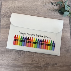 May include: A white envelope-style pouch with a flap closure. The front features a colourful crayon border and the text "Finley's Nursery Master Pieces" in black. The pouch is set against a wooden surface.