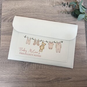May include: A pale-coloured pregnancy notes folder with a flap. The front has a clothesline design with baby clothes and a teddy bear. The text reads "Baby Mclan's Pregnancy Notes".
