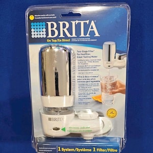 Op de afbeelding: Een verpakt Brita On Tap/En Direct kraanfiltersysteem. Het systeem bevat een filter in twee fasen voor gezonder, lekker smakend water. De verpakking toont het Brita-logo en tekst in het Engels en Frans. Het systeem heeft een elektronische indicator.