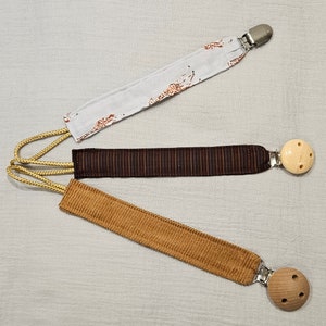 Puede incluir: Tres mordedores de tela para bebé con cuentas de madera y clips metálicos. Los mordedores son marrones, beige y blancos con un estampado de jirafa.