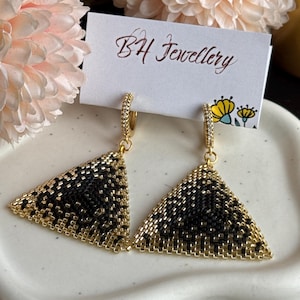 May include: A pair of gold triangle earrings with black and gold beading. The earrings are hanging from gold hoops with clear cubic zirconia stones.