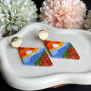 May include: A pair of colorful geometric beaded earrings with gold-toned posts. The earrings are made of small, colorful beads in a diamond shape. The beads are arranged in a pattern of red, orange, yellow, white, blue, and green.