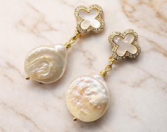 May include: A pair of gold-toned earrings featuring a four-leaf clover design at the top, encrusted with small clear stones. Each earring has a large, iridescent, baroque pearl drop. The earrings are displayed on a marble surface.