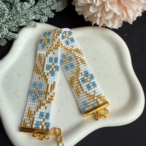 May include: A beaded bracelet featuring a geometric design in white, gold, and pale blue. The bracelet has gold-coloured metal clasps and a chain extender. It is displayed on a white, oval-shaped dish.