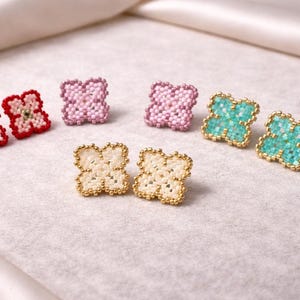Peut inclure: Boucles d'oreilles à clous perlées en forme de trèfles à quatre feuilles. Les couleurs incluent le rouge, le rose, le turquoise et le crème, avec des détails dorés. Présentées sur une surface claire et texturée.
