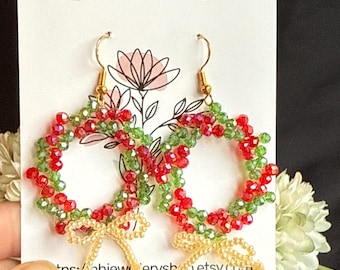 May include: A pair of red and green beaded Christmas wreath earrings with gold bows. The earrings are on a white card with the text "BH Jewellery" and a website address.