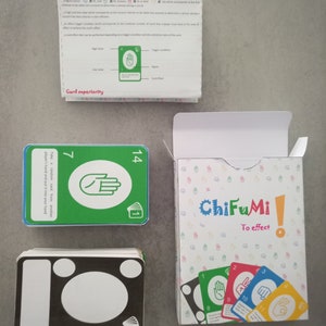 Peut inclure: Un jeu de cartes vert et bleu appelé "ChiFuMi! To effect" avec une carte verte montrant un geste de la main et le numéro 7, une carte verte et bleue montrant un geste de la main et le numéro 14, une pile de cartes noires avec des cercles blancs et une boîte avec le nom du jeu et quelques cartes montrant des gestes de la main.