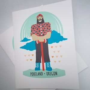 May include: Greeting card featuring a cartoon lumberjack with a red and black plaid shirt, holding an axe. The illustration includes a teal sky, blue clouds, and the text "PORTLAND • OREGON" at the bottom.