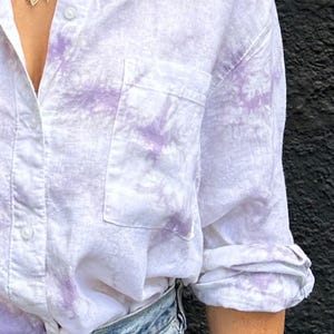 May include: A long-sleeved button-down shirt featuring a white base with a light purple tie-dye pattern, a single chest pocket, and a classic collar. The shirt is paired with light blue denim jeans, and the sleeves are rolled up to the elbows.