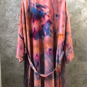 May include: A long, flowing robe with a tie-waist, dyed in shades of pink, blue, and purple. The robe is hanging on a wooden hanger against a textured gray wall. The fabric appears soft and lightweight.