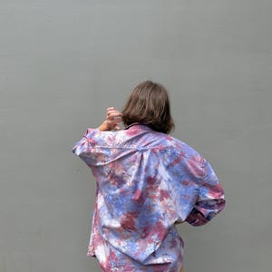May include: A long-sleeved button-down shirt featuring a tie-dye pattern in shades of blue, purple, and reddish-pink. The shirt has a classic collar, a back yoke, and a loose, relaxed fit. It is paired with solid tan-colored trousers.