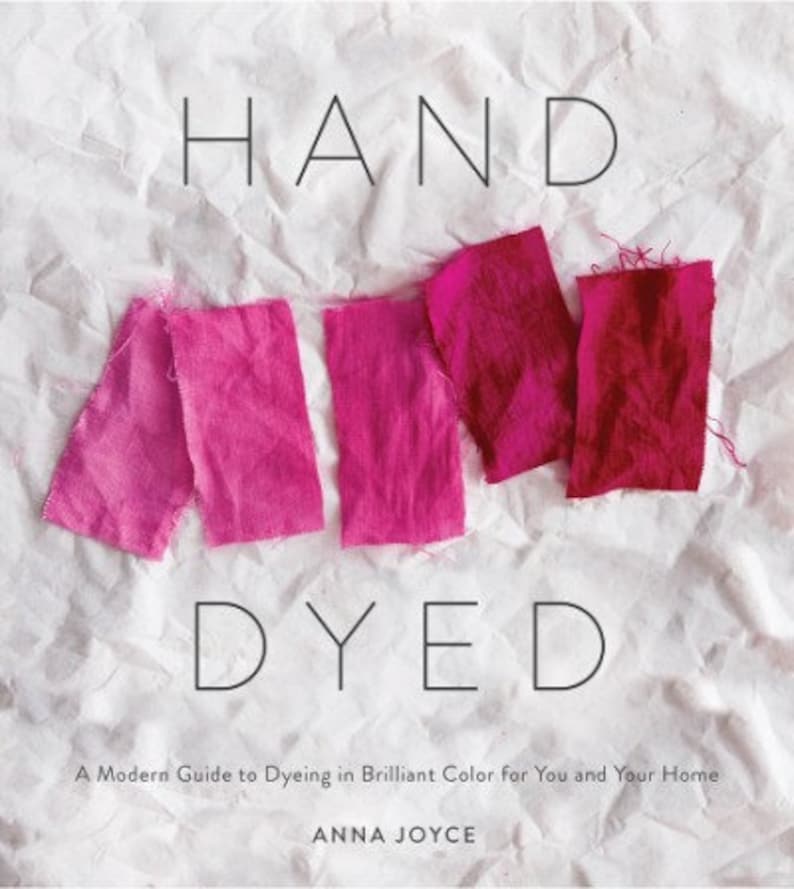 Może przedstawiać: Książka zatytułowana "HAND DYED" z podtytułem "A Modern Guide to Dyeing in Brilliant Color for You and Your Home" autorstwa Anny Joyce. Okładka przedstawia pr&oacute;bki tkaniny w kolorze r&oacute;żowym i czerwonym.