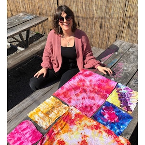 Może przedstawiać: Kobieta w okularach przeciwsłonecznych siedzi przy drewnianym stole piknikowym z r&oacute;żnymi kwadratami tkaniny w stylu tie-dye w odcieniach r&oacute;żowego, ż&oacute;łtego, pomarańczowego, niebieskiego i fioletowego.