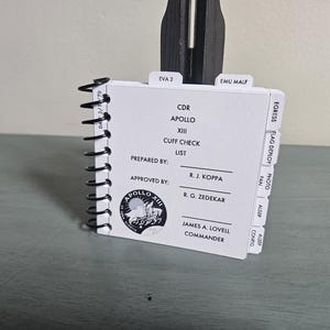 Op de afbeelding: Een spiraalgebonden notitieboek met een witte kaft en zwarte spiraalbinding. De kaft bevat tekst, waaronder "CDR APOLLO XIII CUFF CHECK LIST" en een cirkelvormig logo. Tabs aan de rechterkant zijn gelabeld. Het notitieboek wordt ondersteund door een zwart object.