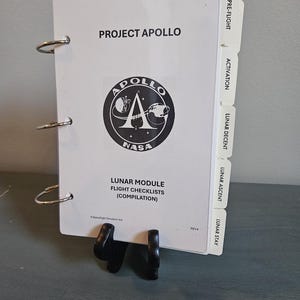 Può includere: Un raccoglitore bianco intitolato "PROJECT APOLLO" con il logo NASA. Contiene "LUNAR MODULE FLIGHT CHECKLISTS (COMPILATION)". Le linguette laterali recitano "PRE-FLIGHT", "ACTIVATION", "LUNAR DECENT", "LUNAR ASCENT" e "LUNAR STAY".