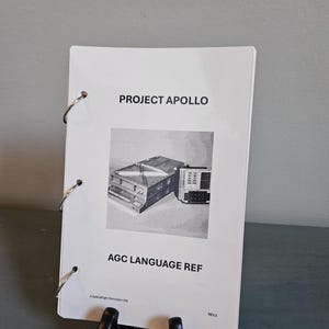 May include: A white booklet titled "PROJECT APOLLO" and "AGC LANGUAGE REF" with a black and white image of a computer. The booklet is held together with metal rings and is propped up by a black stand.