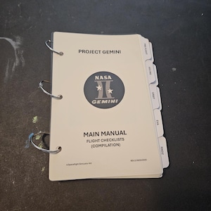 Op de afbeelding: Een witte spiraalgebonden handleiding getiteld "PROJECT GEMINI" met het NASA Gemini-logo. De handleiding bevat vluchtchecklists en is gelabeld "MAIN MANUAL FLIGHT CHECKLISTS (COMPILATION)". De handleiding heeft gelabelde tabbladen voor verschillende vluchtfasen.