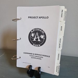 Op de afbeelding: Een witte ringband met de tekst "PROJECT APOLLO" en het NASA-logo. De ringband bevat vluchtchecklists voor de Command & Service Module. Zijkanten tonen secties als "PRE-FLIGHT" en "BOOST".