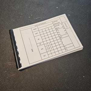 May include: A small, white notepad with a black spiral binding. The pages are filled with a grid-like chart and text, including the words "CATCH UP MANEUVER" and "PURPOSE". The notepad is open on a dark surface.