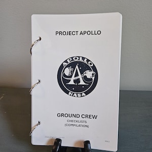 May include: A white booklet titled "PROJECT APOLLO" with the NASA logo and "GROUND CREW CHECKLISTS (COMPILATION)". The booklet is held together with silver metal rings and is propped up by a black stand.