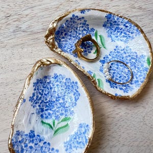 May include: Two white ceramic oyster shell dishes with gold trim and a blue floral pattern. The dishes are decorated with blue hydrangeas and green leaves. One dish has a gold ring and the other has a pearl necklace.
