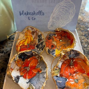 May include: Four handmade oyster shell ornaments with gold trim, featuring colorful floral and pumpkin designs. The ornaments are displayed in a white box, with a card that reads "blakeshells & Co." and an oyster shell illustration.