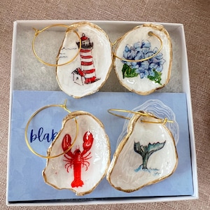 May include: Four pairs of gold hoop earrings with painted oyster shell pendants. The pendants feature a red and white lighthouse, blue hydrangea flowers, a red lobster, and a whale tail. The earrings are presented in a white box with a blue insert.