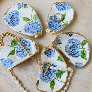 May include: Five white oyster shell ring dishes with blue hydrangea floral designs and gold edges. A gold chain necklace and a pearl necklace are displayed on the dishes. A gold ring is on one dish.