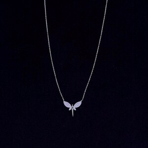 Puede incluir: Un collar de cadena de plata con un colgante de libélula. La libélula tiene alas transparentes con pequeñas piedras brillantes.