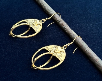 May include: A pair of gold-toned earrings with a detailed design featuring a bird and a circle. The earrings are hanging from a wooden stick.